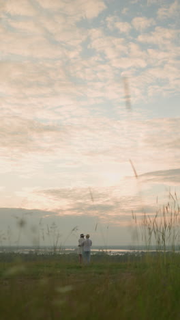 partners stand on a grassy hill beside a lake at sunset, gazing at the sky. the man, in a white shirt, hat, and jeans, lovingly holds the woman by the waist. she wears a black hat and a white dress