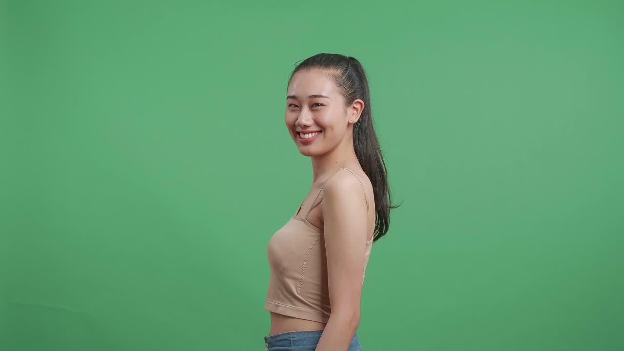 스튜디오의 녹색 스크린 배경에 서서 카메라를 향해 돌아서서 미소 짓는 아시아 여성의 측면 시각