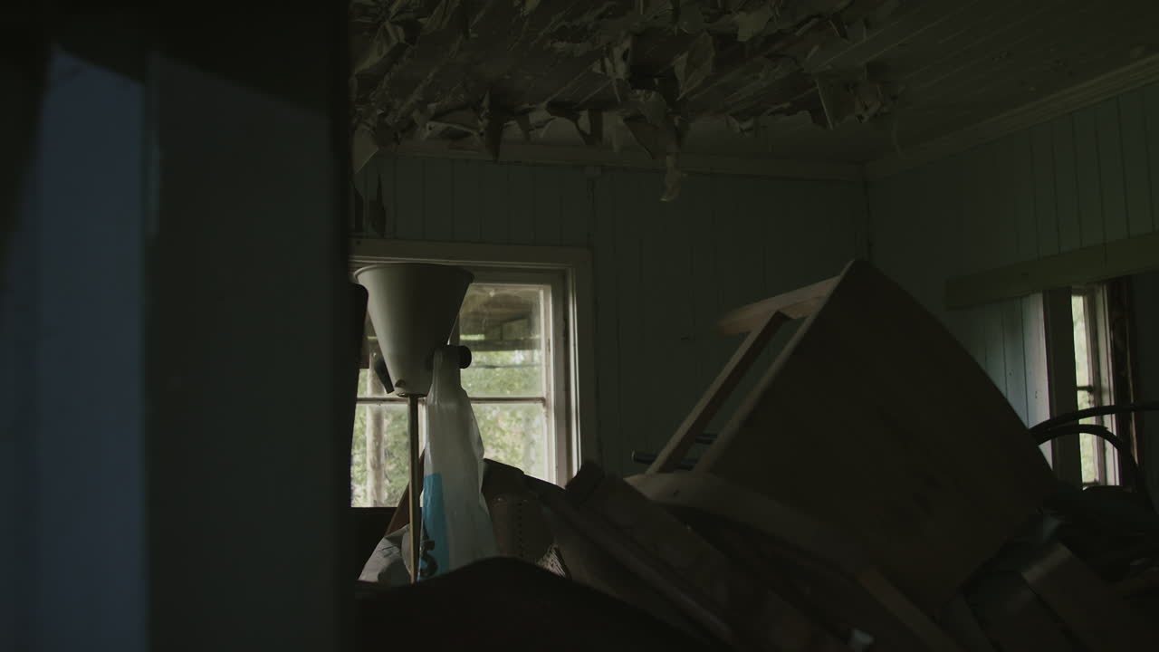 Dimly lit room of a neglected house, showing damaged ceiling and cluttered furniture.