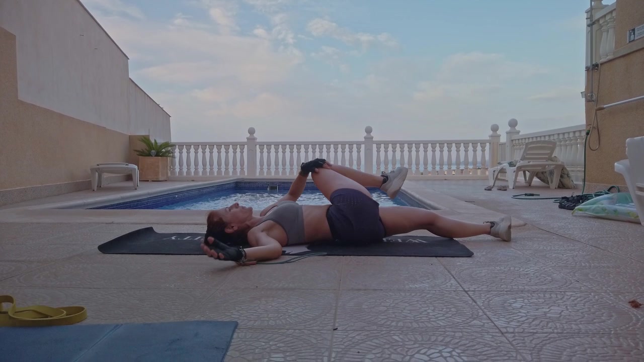 Inquadratura dal basso di una donna che esegue allungamenti dei glutei con ginocchio al petto su un tappetino nero accanto a una piscina su una terrazza. Routine di mobilità e defaticamento all'aperto