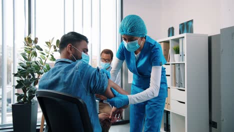 Rückseite-Der-Hinduistischen-Jungen-Weiblichen-Krankenschwester-In-Uniform,-Die-Einem-Männlichen-Patienten,-Der-Im-Kabinett-Im-Krankenhaus-Sitzt,-Eine-Coronavirus-Impfstoffinjektion-Vornimmt,-Während-Der-Arzt-Dahinter-Arbeitet,-Covid-Impfkonzept