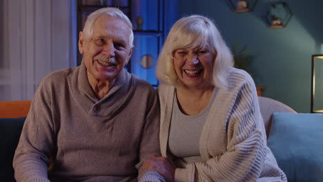 Happy-old-senior-elderly-family-couple-hugging,-laughing,-smiling-looking-at-camera-at-home-sofa