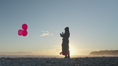 La-Silueta-De-Una-Mujer-Sostiene-Tres-Globos-Rojos-En-La-Playa-Mientras-Se-Pone-El-Sol