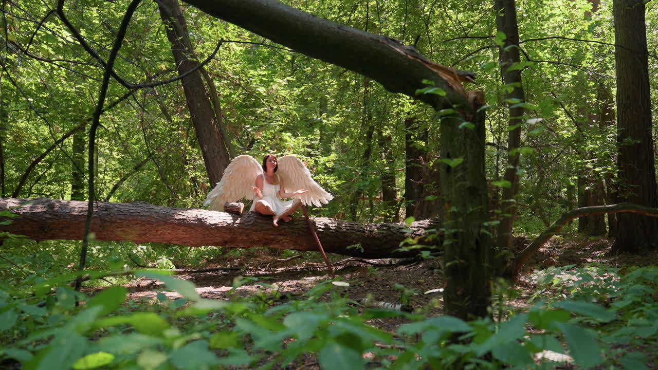Woman dressed in white sits cross legged on fallen forest log practicing yoga in lush green woodland, surrounded by sunlight and trees, with wooden staff and hand drum placed beside her
