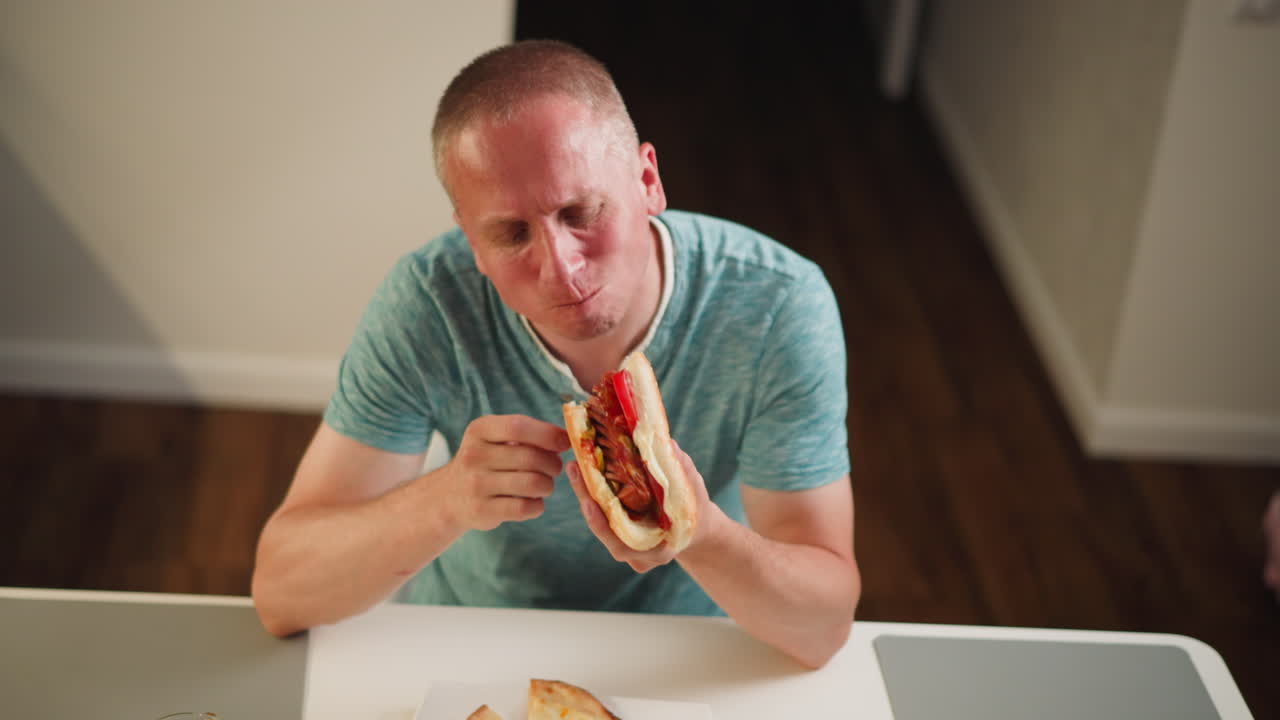 Top view of man sitting at table enjoying big burger, savoring every bite, wearing casual greenish blue shirt, relaxed at home, eating juicy sandwich with tomatoes