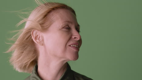 Close-Up-View-Of-Blonde-Mature-Woman-Wearing-Green-Shirt-Posing-At-Camera-Touching-Her-Hair-While-The-Wind-Move-Her-Hair-And-Shirt-On-Green-Background