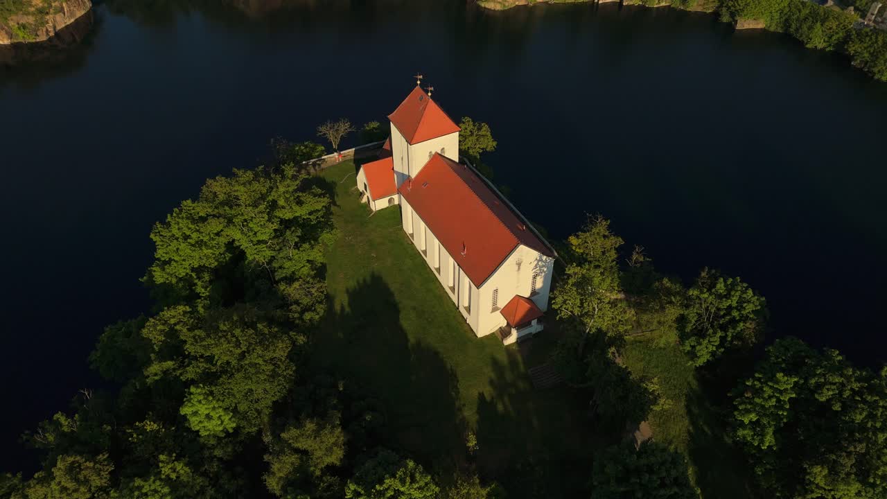 Drone view of the historic Wehrkirche Beucha in Saxony, Germany. The church stands atop a cliff surrounded by a flooded quarry, with the evening sun casting warm light across the landscape.