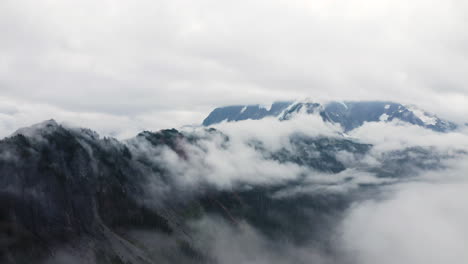 Drone-ascends-above-fluffy-white-clouds-as-they-rise-slow-above-mountain-ridgeline-and-obscure-peak