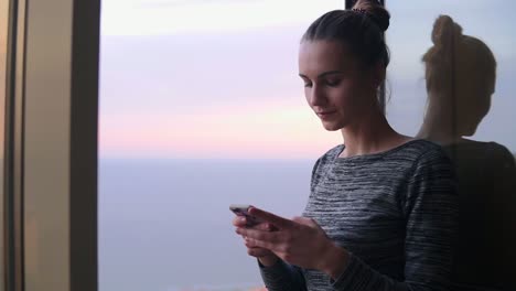 Vista-De-Cerca-De-Una-Hermosa-Joven-Sentada-Junto-A-La-Ventana-Abierta-Con-Una-Sonrisa-Mirando-El-Teléfono-Y-Escribiendo-Un-Mensaje-Durante-La-Puesta-De-Sol