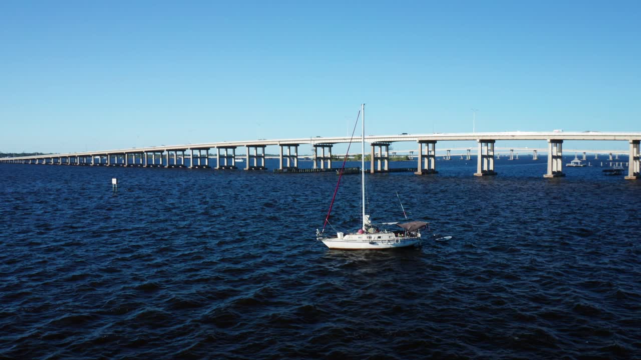Una barca a vela riposa su acque di un blu profondo vicino a un lungo ponte a Fort Myers, Florida. La scena cattura una giornata calma e soleggiata, perfetta per video di viaggio o temi marittimi