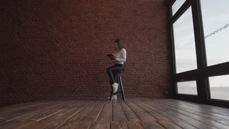 Female-sit-on-high-seat-in-empty-room-near-bright-window-and-red-brick-wall