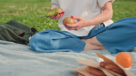 lower view of sisters on picnic blanket sharing fruit and sandwich, one taking orange from canvas bag and giving to other, hands closeup on green grass during relaxed summer outdoor sibling bonding