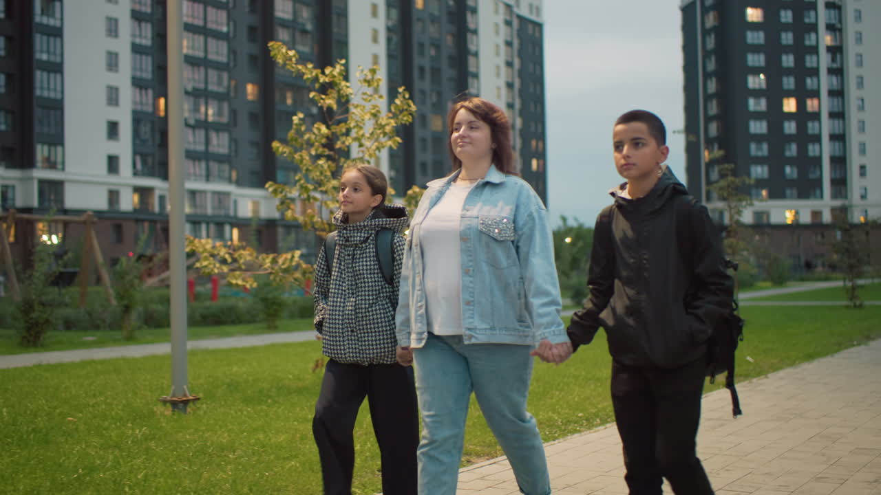 Woman walks hand in hand with two children through green urban park area near modern apartment buildings during evening, showing warmth, care, and connection in everyday city life with school backpacks