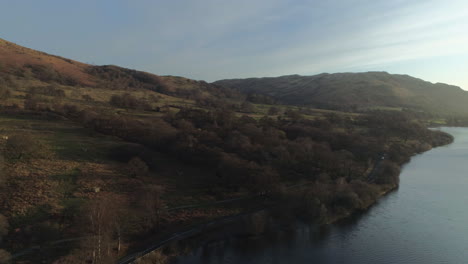 Luftdrohne-Bei-Sonnenaufgang-über-Dem-Ullswater-See-Bei-Sonnenaufgang-Mit-Hügeln-Und-Bäumen-Und-Einem-Auto,-Das-Am-Lake-District-Von-Cumbria,-Großbritannien,-Vorbeifährt