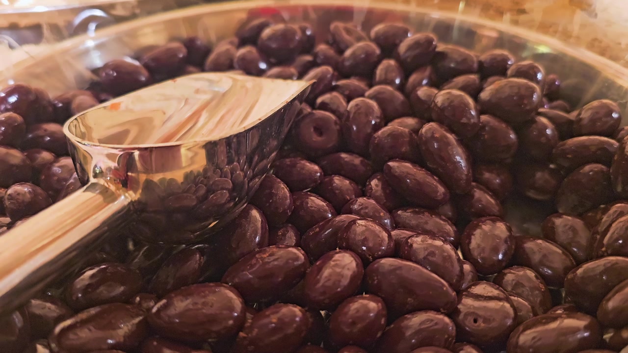 Detailed snack counter shot of chocolate coated nuts piled high in a clear tray, with serving scoop reflections, warm retail lighting, and tempting bulk candy shop texture.