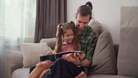 A-little-brunette-girl-in-a-pink-dress-sits-on-the-lap-of-her-brunette-male-father-in-a-green-checkered-shirt-and-tunes-the-ukulele-to-play-a-musical-instrument-while-sitting-on-a-gray-sofa-at-home-in-a-modern-apartment