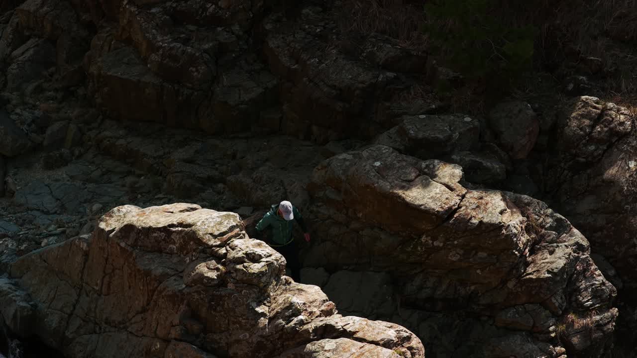Lone hiker with white hat walking on rock formation along pristine stream, rugged mountain landscape, Praglia. Italy. Aerial drone top-down view, low altitude