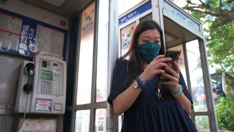 low-angle view of a woman uses a smartphone next to public telephone booths, also called a payphone, as they are rapidly dying out and disappearing in the age of smartphones and the internet