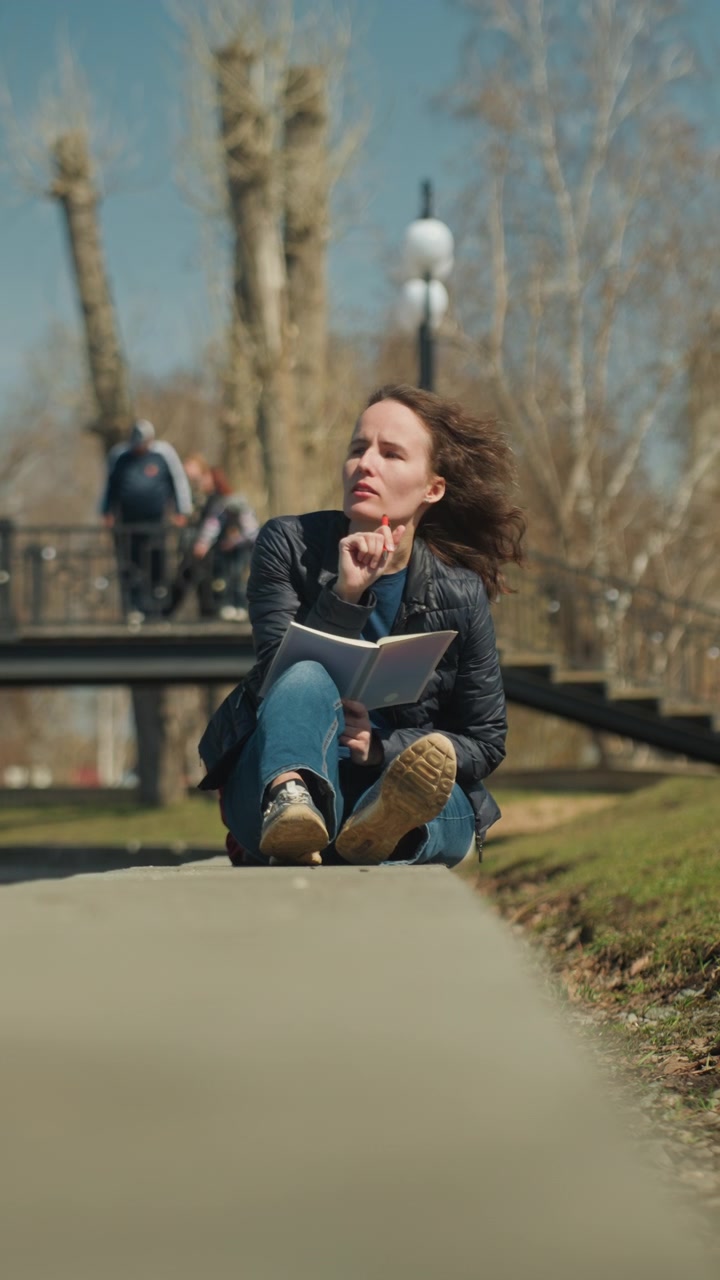 Primo piano di una donna che legge un libro all'aperto in una giornata di sole. Il libro è tenuto tra le mani mentre è seduta a gambe incrociate, con il vento che le soffia delicatamente tra i capelli. L'ambientazione è un parco con un ponte, alberi e persone che camminano sullo sfondo.