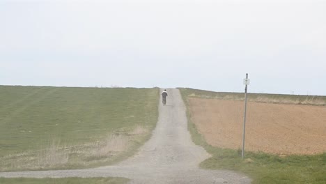 a cyclist pedalling along a dusty gravel road that leads through two fields on a cloudy day