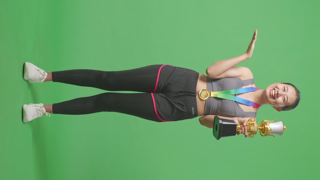 cuerpo lleno de mujer asiática con una medalla de oro mirando un trofeo de oro en sus manos, sonriendo, y señalando a un lado en el fondo de la pantalla verde en el estudio