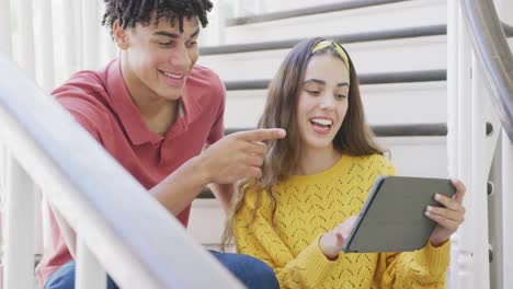 pareja biracial feliz pasando el tiempo en casa usando tableta y sentada en las escaleras