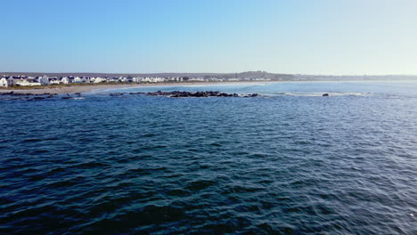 Drone-approach-over-sea-towards-beach-lined-with-whitewashed-houses,-Paternoster