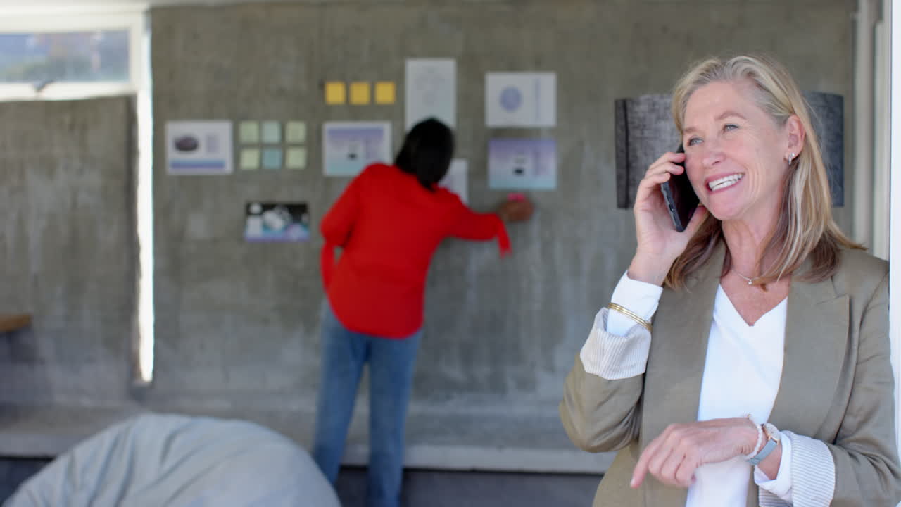 Talking on smartphone, senior woman smiling while colleague working on wall display, in office