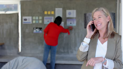 talking on smartphone, senior woman smiling while colleague working on wall display, in office