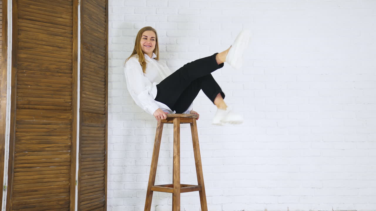 Smiling long-haired lady waving her feet sitting on a high stool. Model demonstrating white loafers with massive soles at white wall backdrop.