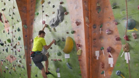 Male-climber-climbs-the-cliff-wall-in-the-room-reaching-and-gripping-hold.-A-professional-concentrated-climber-moves-up-the-wall-the-camera-rises-and-lays-down-his-movements.-Boom-and-truck-follow-camera.