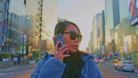 beautiful asian oriental female girl woman model in big city town urban street area with sunglasses and cellphone talking and standing near skyscrapers