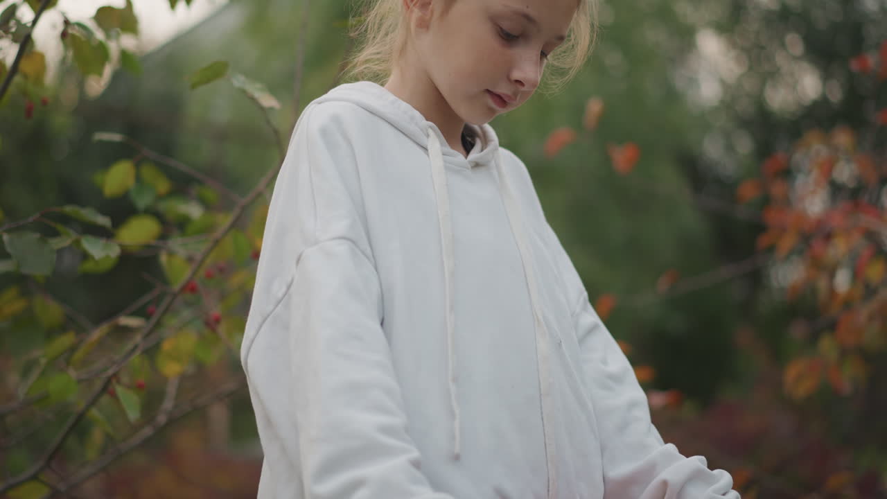 Junges Mädchen hält Apfelschale und trägt einen weißen Kapuzenpullover, geflochtenes Haar. Holzschale gefüllt mit reifen Äpfeln, Hintergrund eines belaubten Obstgartens, warmes Herbstlicht, nachdenklicher Ausdruck, Ernteaktivität im Freien.