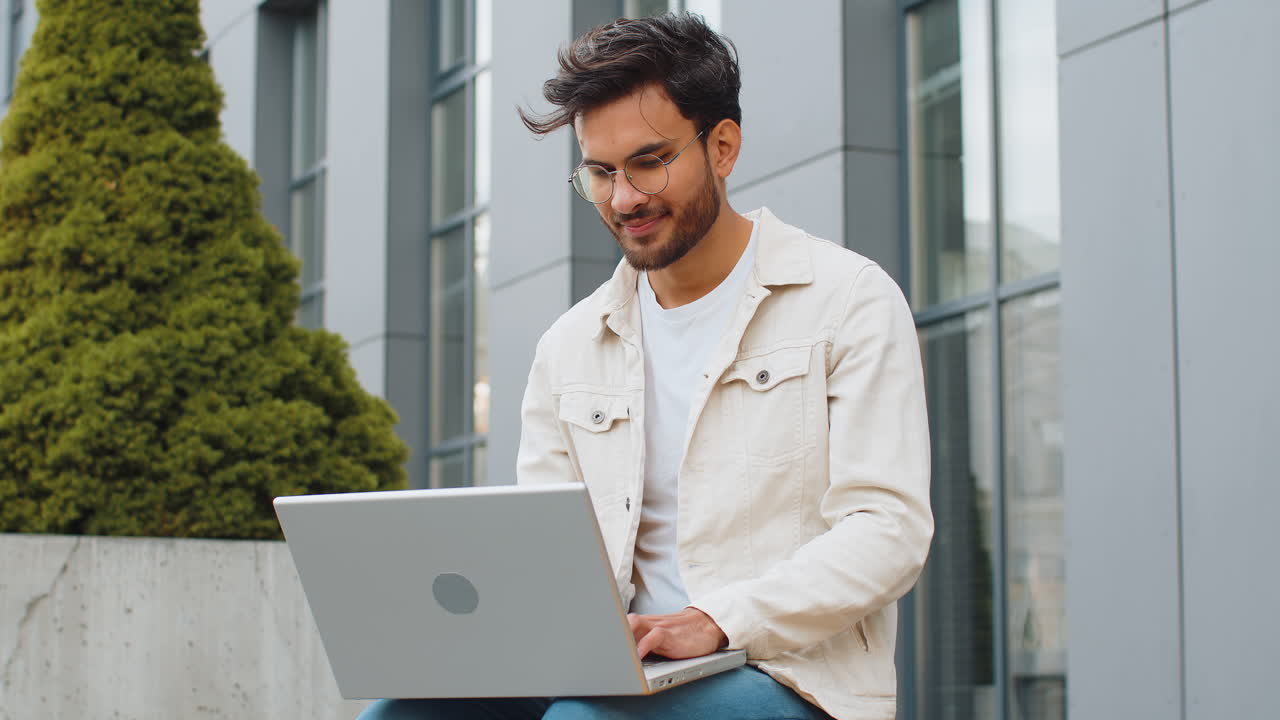 Indian man freelancer working on laptop computer sends messages reading email outdoors on bench