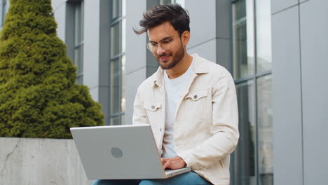 indian man freelancer working on laptop computer sends messages reading email outdoors on bench