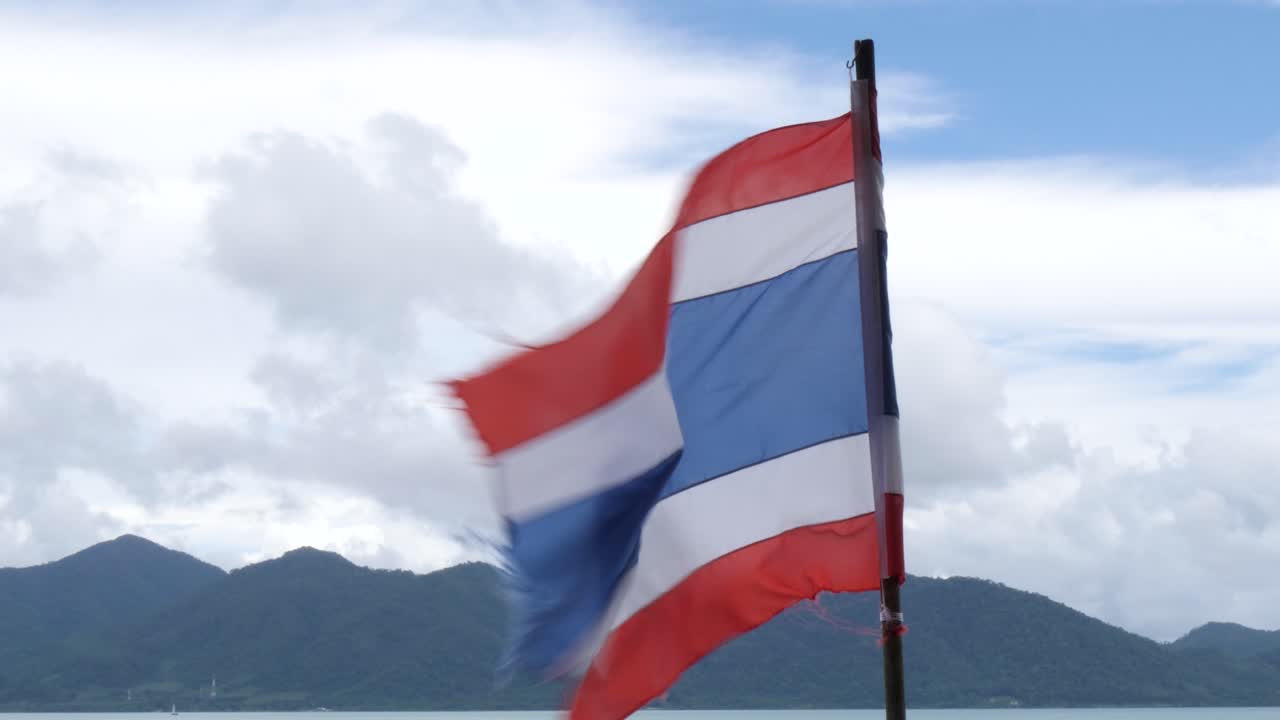 A torn Thai flag blows in the wind against mountain and ocean views in Thailand, showing weathered fabric and strong coastal breeze in a scenic shoreline setting