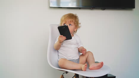 little child on white t-shirt plays with phone on chair