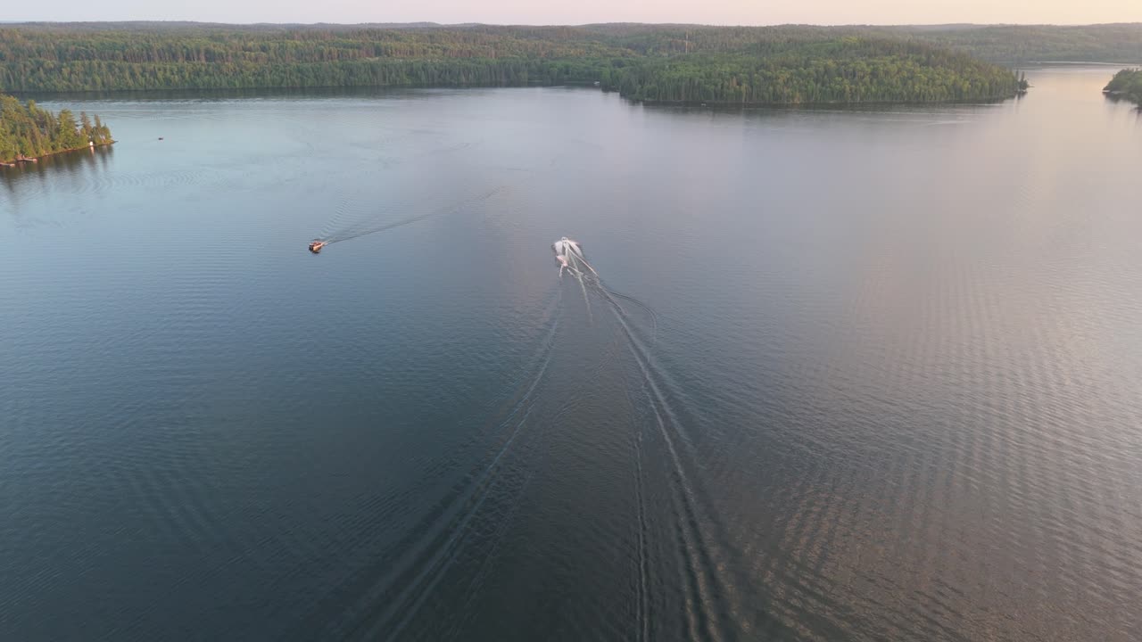 Aerial drone footage of boats moving across a calm lake, leaving wake trails behind in the peaceful water at sunset