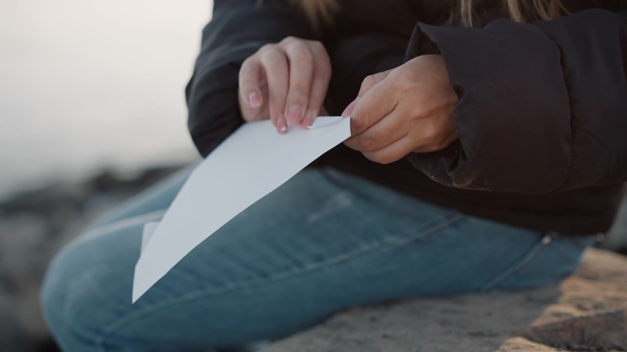 Woman sits outdoors on rock holding and examining white paper. Woman wears blue jeans and dark jacket with neatly manicured nails. background is softly blurred suggesting open space