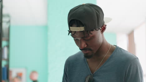 close portrait of young man in cap looking down, then turning toward blurred people in background, thoughtful expression in indoor light, teal wall and shelves