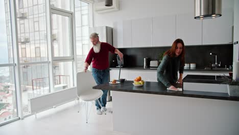 An-elderly-man-with-gray-hair-and-a-lush-beard-in-a-red-T-shirt-is-cleaning-his-home-with-his-adult-brunette-daughter-in-a-green-sweater-in-a-modern-kitchen