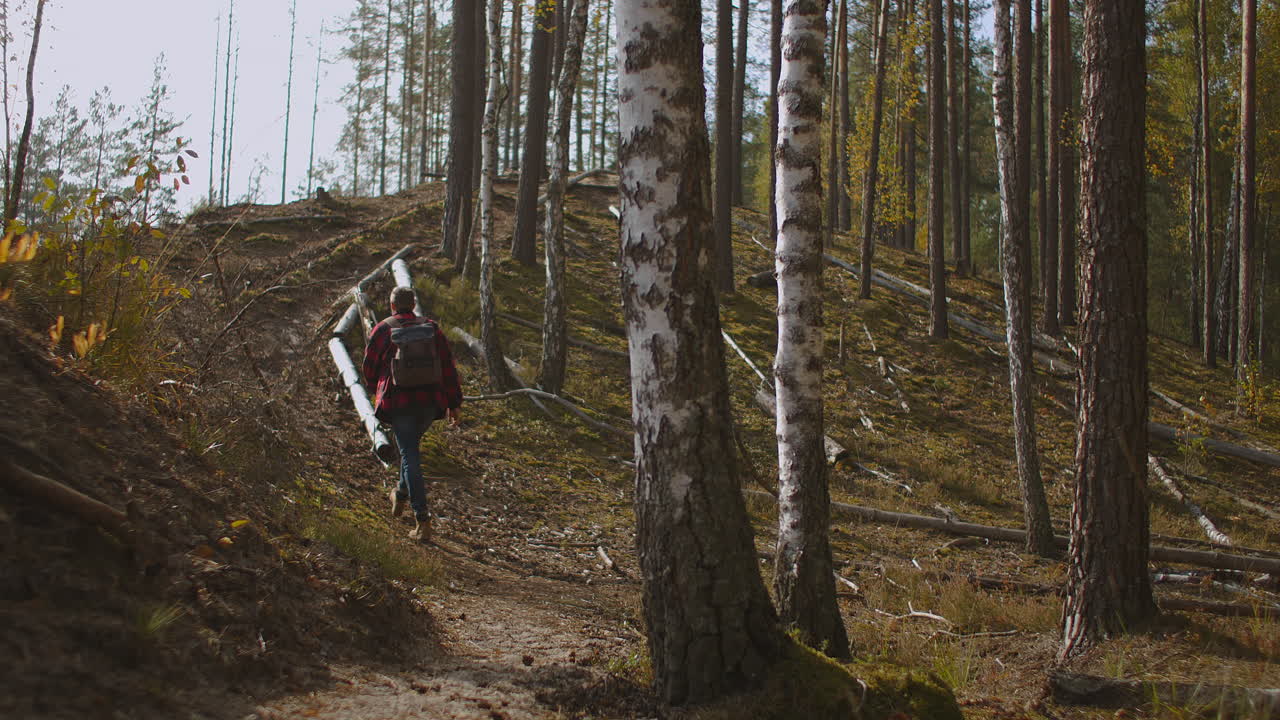 Young man with backpack walking on the hill against the mountains at sunset in autumn. Landscape with sporty guy meadow snowy rocks orange trees houses. High quality 4k footage