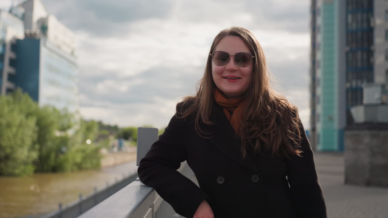 Stylish woman stands near city river leaning on safety railing as mood shifts from crying to laughing, sunlight touching her hair while breeze flows through urban scenery creating emotional contrast