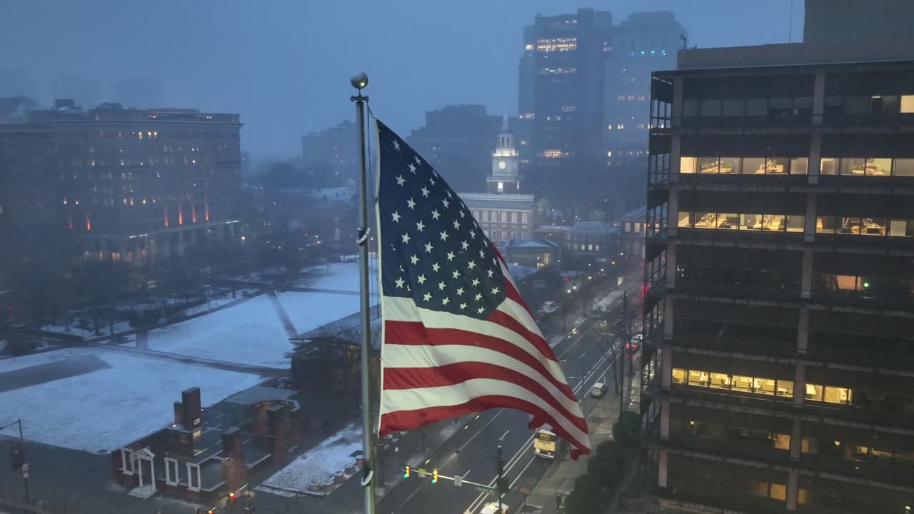 Vista aerea innevata di una grande bandiera americana che sventola sopra il centro di Philadelphia, con strade invernali, edifici storici e luci della città. Atmosfera urbana patriottica e malinconica negli Stati Uniti. Primo piano