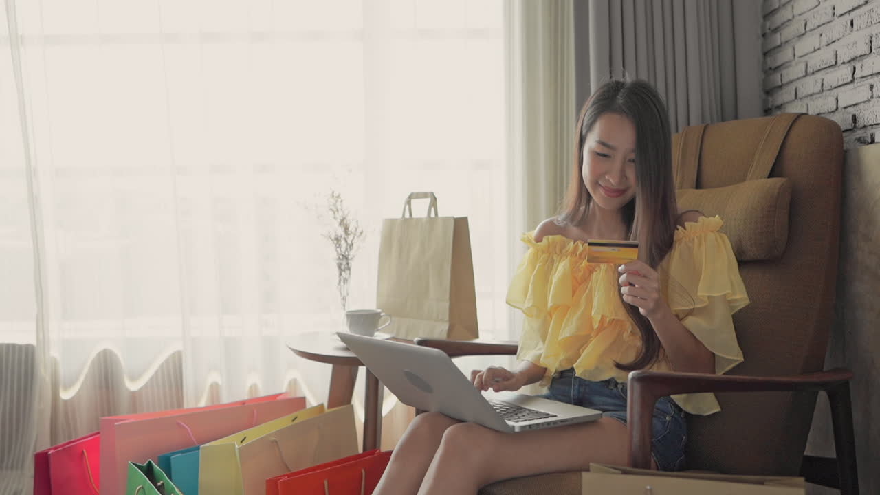 A young attractive woman sitting in a big comfortable easy chair and surrounded by shopping bags, types her credit card number into her laptop.copy space