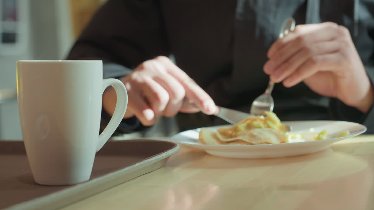 Mug plate eggs during breakfast, soft sunlight on wooden table, hands using fork and knife to cut into yolk, steam rising from coffee mug, intimate slow morning ritual in cozy cafe setting, casual