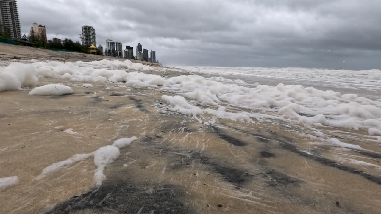 모래를 고 있는 바다 거품으로 폭풍이 불고 있는 해변 장면, 구름이 가득한 하늘 아래, 자연의 원시적인 힘과 오염을 보여준다.