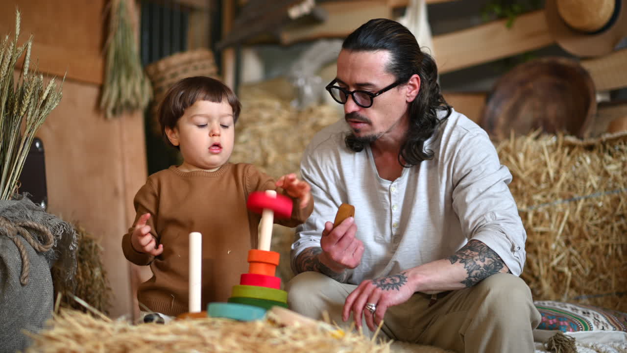 Father playing with his son with colourful, ecological wooden toys in a barn, near square hay bales