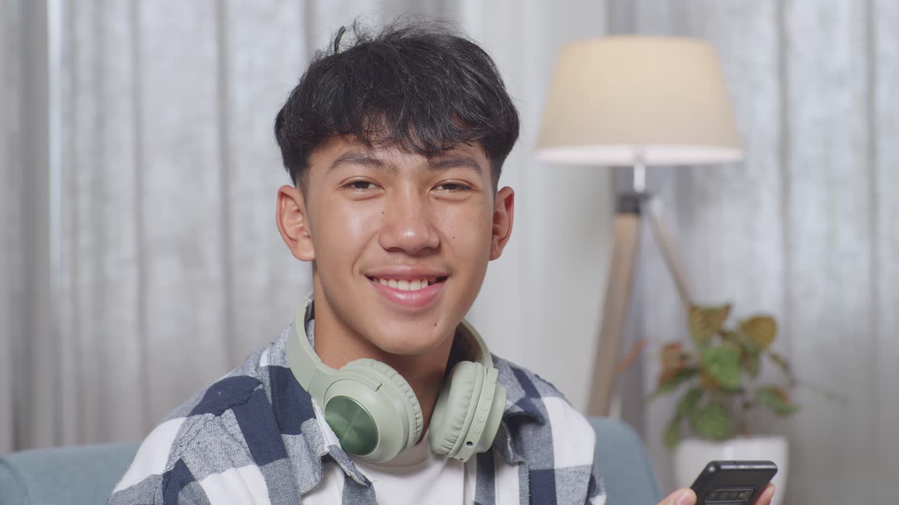 가정의 거실 소파에서 스마트폰을 재생하는 동안 헤드폰을 들고 카메라를 향해 미소 짓는 아시아 십대 소년의 클로즈업