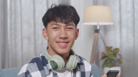 close up of asian teen boy with headphones smiling to camera while playing smartphone on sofa in the living room at home